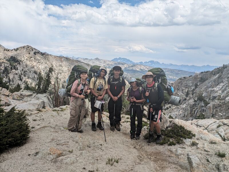 A group of four backpackers are carefully crossing a shallow, rocky stream in a forest. They are heavily laden with gear, including sleeping bags and backpacks. The water is clear, and the streambed is covered with colorful rocks. The surrounding forest is lush and green, with trees lining the banks of the stream.